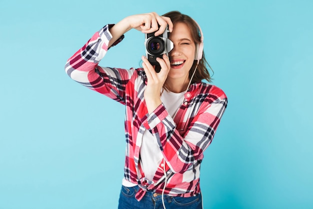 Portrait of young lady in headphones looking in little retro camera while taking photo on over pink background