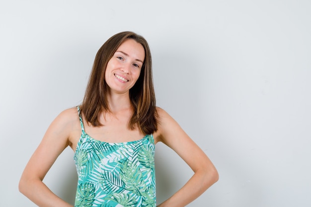 Portrait of young lady keeping hands on hip in blouse and looking cheerful front view