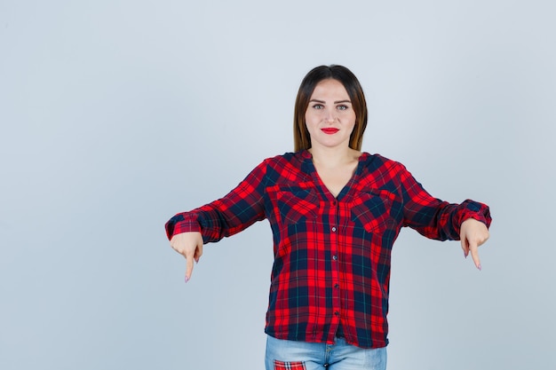 Portrait of young lady pointing down in checked shirt, jeans and looking confident front view