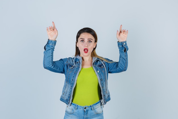 Portrait of young lady pointing up in denim outfit and looking surprised front view