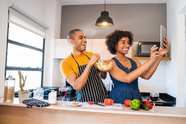 Portrait of young latin couple cooking together and taking a selfie with digital tablet in the kitchen at home