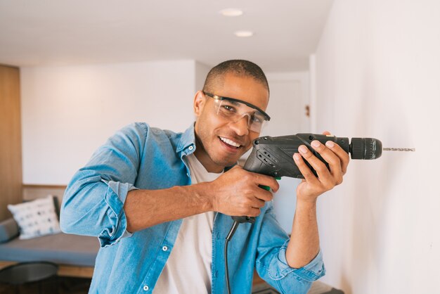 Ritratto di giovane uomo latino con un trapano elettrico e fare un buco nel muro. interior design e concetto di ristrutturazione della casa.