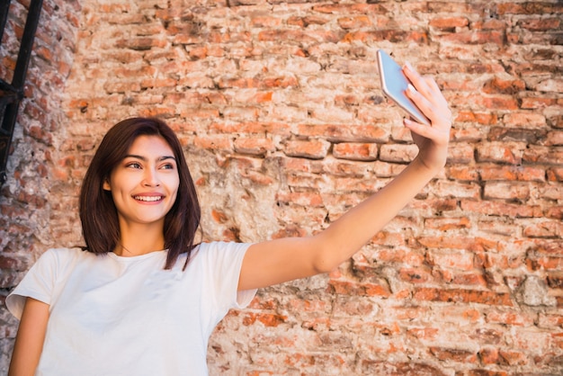 벽돌 벽에 야외에서 그녀의 mophile 전화 selfies를 복용하는 젊은 라틴 여자의 초상화. 도시 개념.