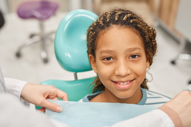 Портрет молодой маленькой девочки, улыбающейся спереди, сидя на стуле, она посещает стоматолога