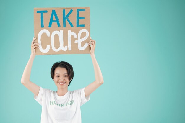 Portrait of young lovely Asian woman holding placard with take care inscription above hr head and smiling at camera