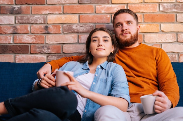 Portrait of young loving couple hugging, holding cups of warm drink and watching romantic movie on TV at evening together sitting on comfortable sofa in cozy living room. Concept of household life.