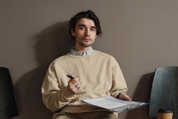 Portrait of young male candidate looking at camera while\
filling application form for getting job on chair in corridor