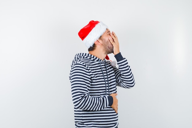portrait of young male holding hand on mouth in hoodie, santa hat and looking depressed 
