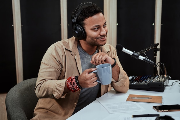 Portrait of young male radio host going live on air talking\
with guest holding a cup of coffee or