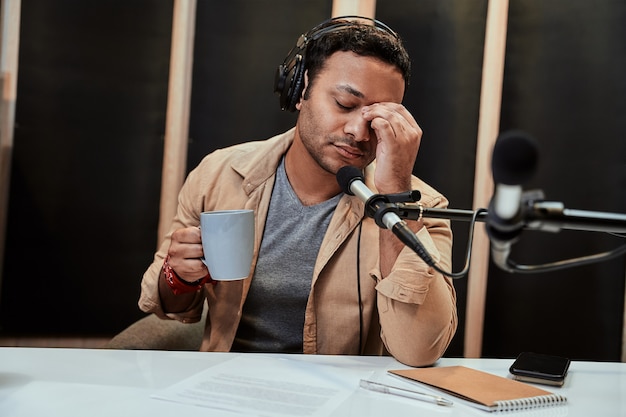 Ritratto di un giovane conduttore radiofonico maschio in cuffia che sembra stanco esausto che beve caffè mentre