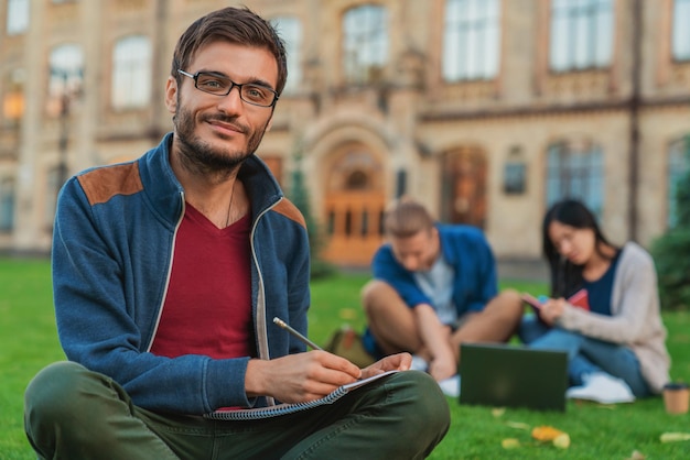 Ritratto di giovane studente maschio che nota mentre gli studenti utilizzano il laptop in background al prato