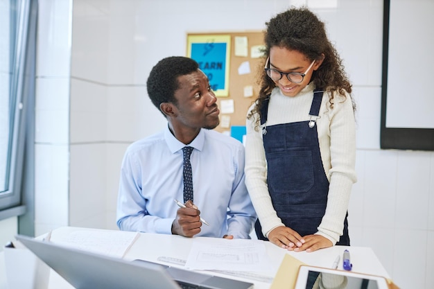 Portrait of young male teacher smiling while working with african american girl in school classroom