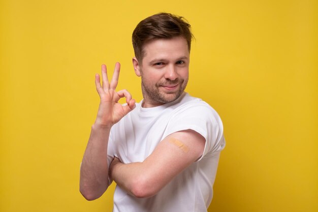 Portrait of young man after getting receive anti virus vaccine covid19 Showing arm and ok gesture Studio shot on yellow wall