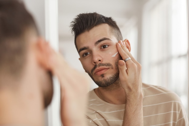 Ritratto di giovane che applica la crema per il viso mentre si guarda allo specchio durante la routine mattutina per la cura della pelle, copia spazio