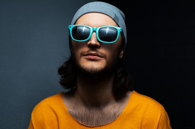 Portrait of young man on black and grey background, wearing blue sunglasses, hat and orange shirt.