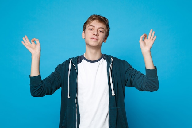 Portrait of young man in casual clothes holding hands in yoga gesture, relaxing meditating isolated on blue wall. 