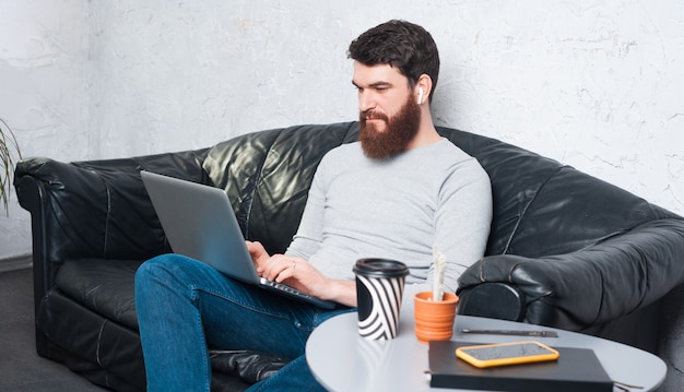 Портрет молодого человека в случайных сидя на диване и работает на ноутбуке