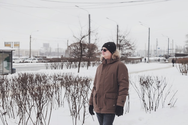 Портрет молодого человека в повседневной зимней одежде в очках на прогулке по городу. Привлекательный подросток стоит в старом городе, серьезно смотрит в сторону, но слегка улыбается в морозный пасмурный день. Копировать пространство