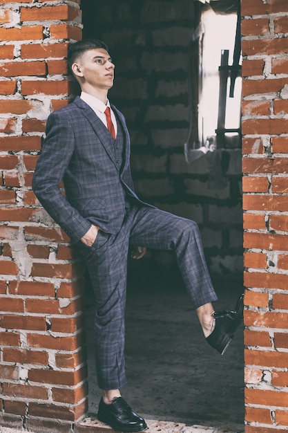 Portrait of young man in classic suit and black patent leather shoes leaning in the doorway of brown brick wall, toned