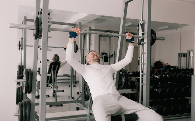 Ritratto di un giovane uomo che fa esercizi fisici, con un allenamento del viso teso con un bilanciere seduto su una panchina in palestra. in una maglietta bianca. uno stile di vita sano