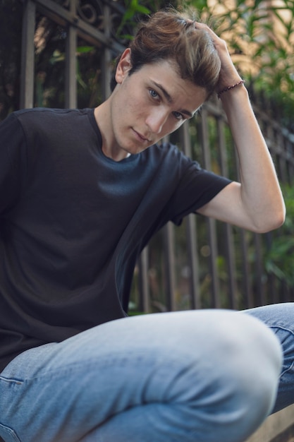 Фото Портрет молодой человек, одетый в черный сарай и джинсы. сидеть на заборе и смотреть неподвижно.
