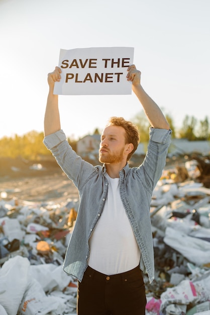 Portrait of young man fighting for nature holding save mother earth sign at dump. Protesting against nature pollution waving hands calling on to save planet.