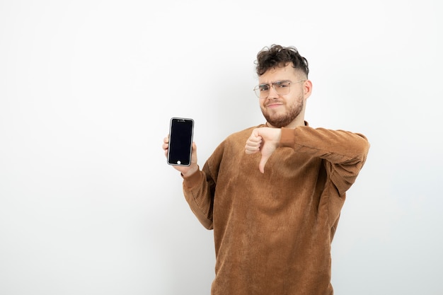 Portrait of young man in glasses holding cellphone and giving thumbs down. 