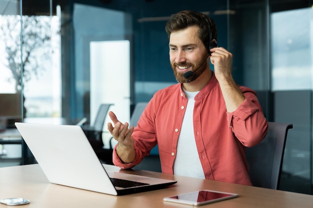 Portrait of a young man in a headset and a red shirt he sits at the desk in the office works on a