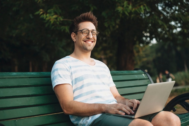 Portrait of a young man looking at camera smiling while holding a laptop on his legs outside in the park.