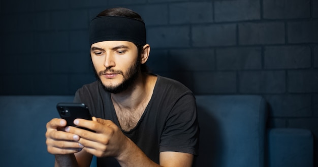 Portrait of young man looking in smartphone, sitting on sofa on background of black brick wall.