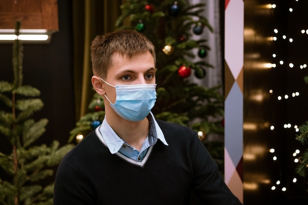 Портрет молодого человека в медицинской маске и свитере на фоне боке и елки