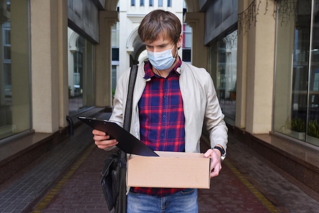 Portrait of a young man in a medical protective mask with a box\
of things who has lost his job