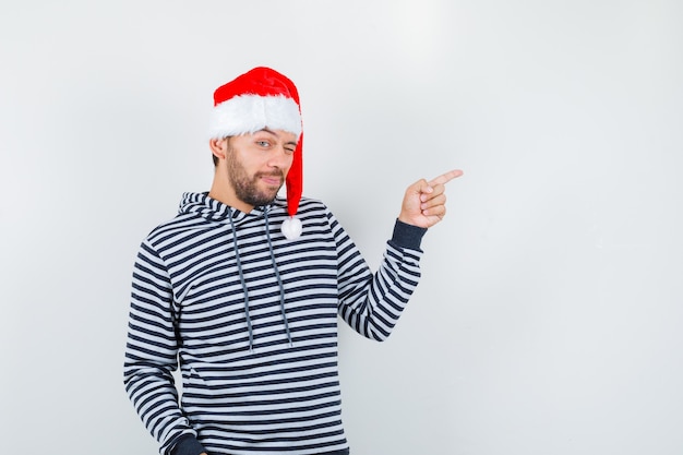 Ritratto di giovane uomo che punta a destra mentre sbatte le palpebre in felpa con cappuccio, cappello di babbo natale e guarda fiducioso vista frontale