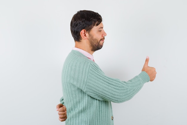Портрет молодого человека, показывающего большой палец вверх в рубашке, кардигане и выглядящего довольным
