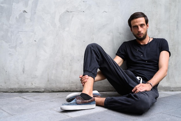 Portrait of young man sitting on footpath against wall