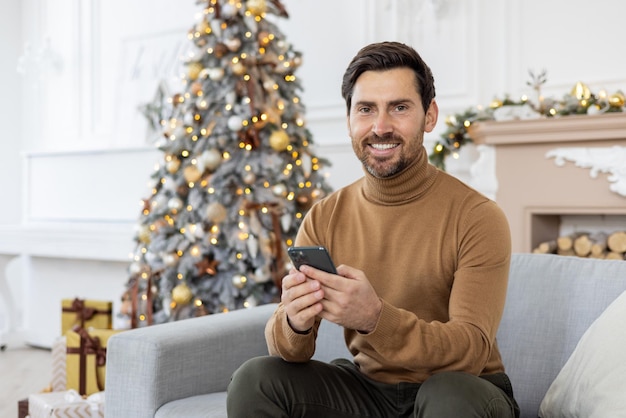 Портрет молодого человека, сидящего дома на диване возле рождественских украшений и рождественской елки