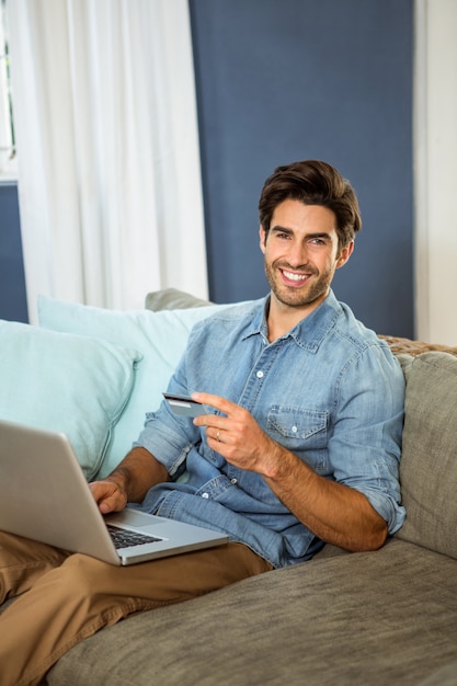 Photo portrait of young man sitting on sofa and shopping online on laptop