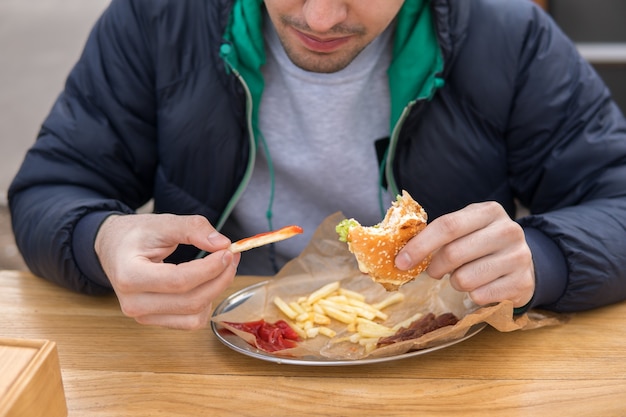 A portrait of young man in street food cafe. He eats french fries with ketchup and burger.