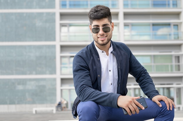 Портрет молодого человека в стильных солнцезащитных очках позирует, сидя на скамейке в городской среде