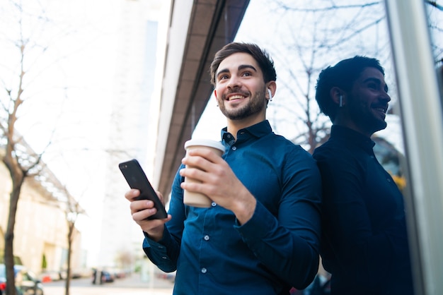 Ritratto di giovane uomo utilizzando il suo telefono cellulare e tenendo una tazza di caffè mentre si trovava all'aperto in strada. concetto urbano.