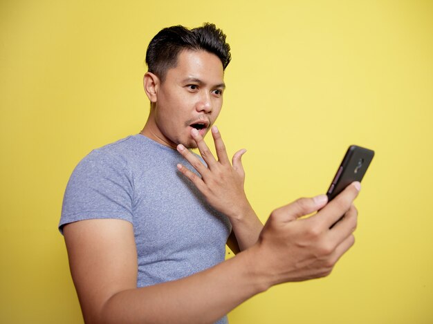 黄色の背景で隔離の携帯電話を見て幸せな肖像画の若い男は非常に驚いた