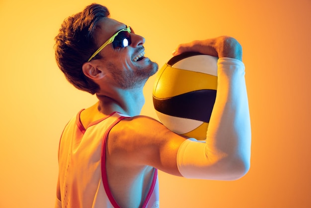 Portrait of young man volleyball player in motion kicking ball playing isolated over yellow studio background in neon light