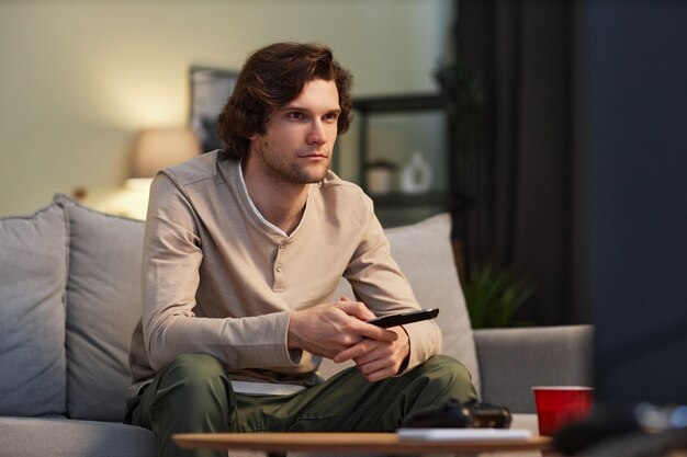 Portrait of young man watching TV at home sitting on couch interior copy space