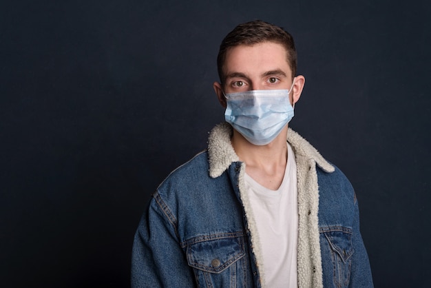 Portrait of a young man wearing jeans jacket in a white medical face mask for personal protection during the COVID19 coronavirus pandemic