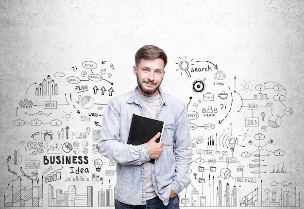 Portrait of a young man wearing a jeans shirt and holding a notebook standing near a concrete wall with a business idea sketch drawn on it.