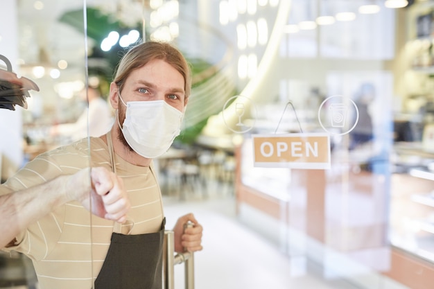 Portrait of young man wearing mask while opening cafe in morning with focus on OPEN sign at glass door, copy space