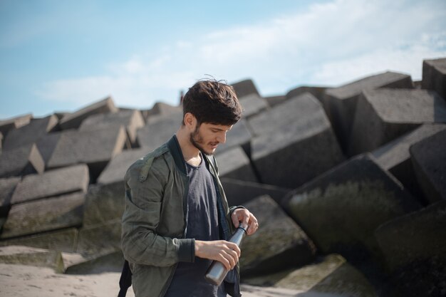 Portrait of young man with backpack, using reusable steel thermo water bottle, outdoor on of square stones.