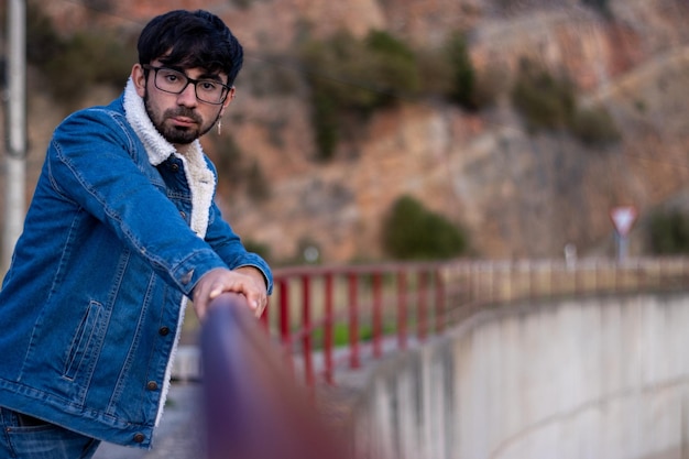 Portrait of a young man with glasses and a beard dressed in a denim jacket leaning on a fence