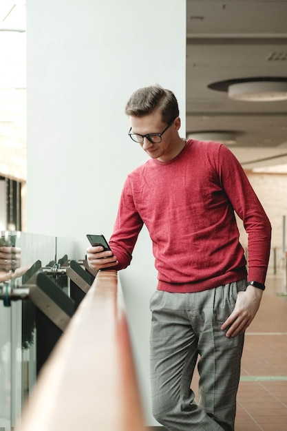 Portrait of a young man with glasses and with a smartphone in his hands near a glass fence in mall