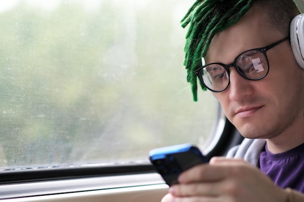 Портрет молодого человека с зелеными дредами в очках, наушниках, слушающего музыку, просматривающего мобильный телефон, едущего в поезде.